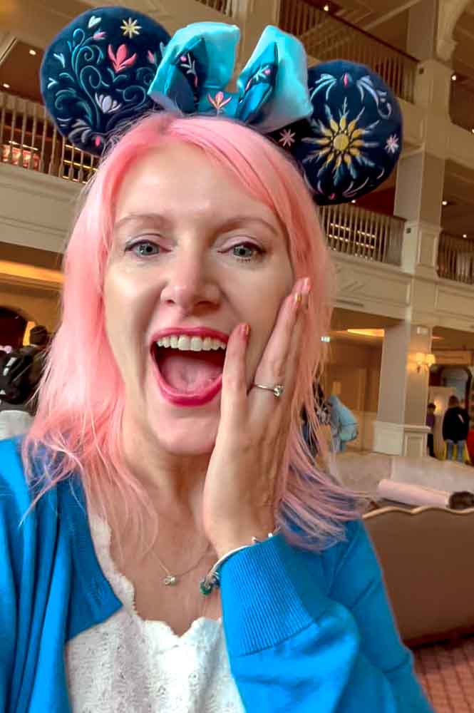 A woman with pink hair and blue mouse ears looks surprised and delighted in the posh lobby of the best Disneyland Paris hotel, one of a few excellent choices for luxury stays or famil-friendly budget hotels the resort has to offer