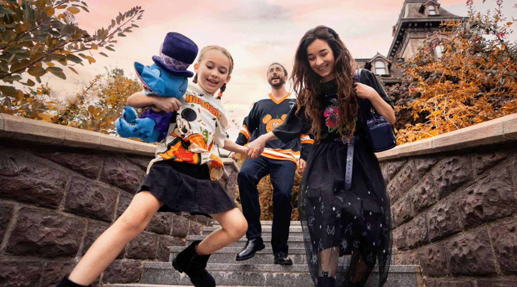 A young family skips down the steps outside Phantom Manor wearing Disney Halloween Festival 2026 merchandise including seasonal inspired tshirts and the little boy is carrying a Halloween Stitch doll