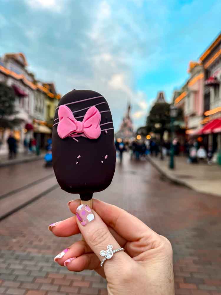 Chocolate-covered Minnie Mouse cake pop decorated with a pink bow, held up in front of Main Street, U.S.A. at Disneyland Paris.