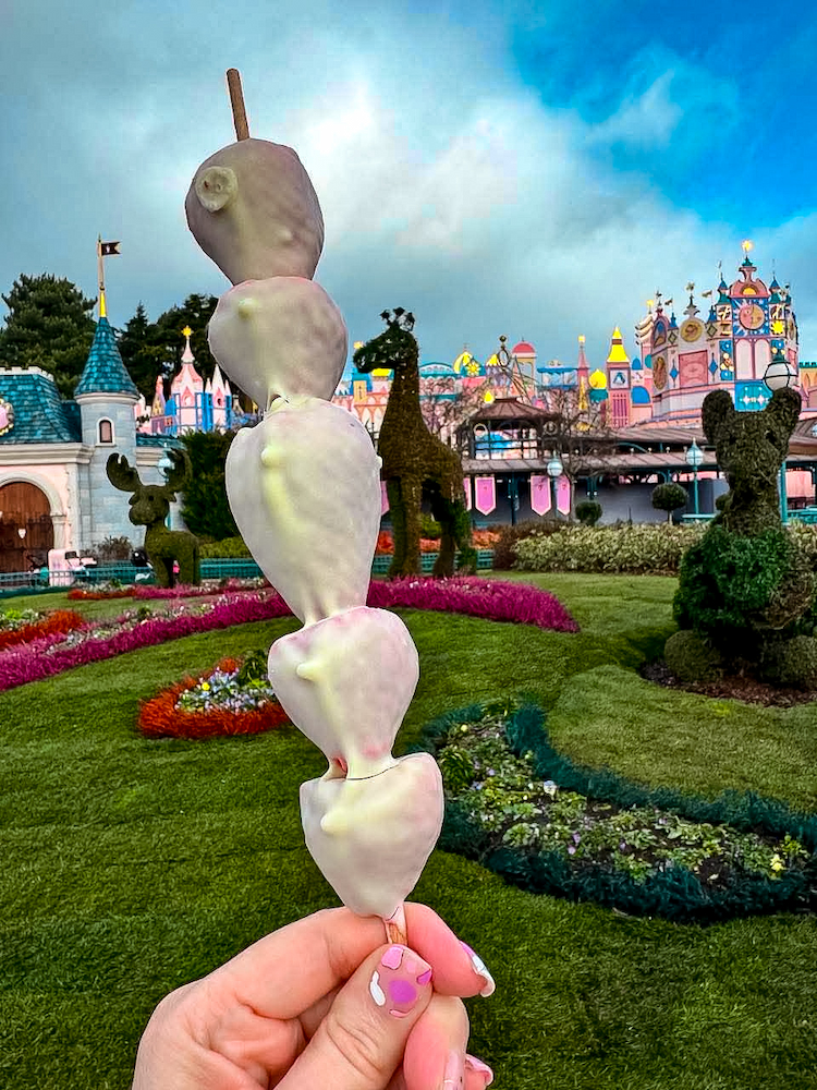 Skewer of five chocolate-coated strawberries held up in front of It's a Small World in Fantasyland at Disneyland Paris.