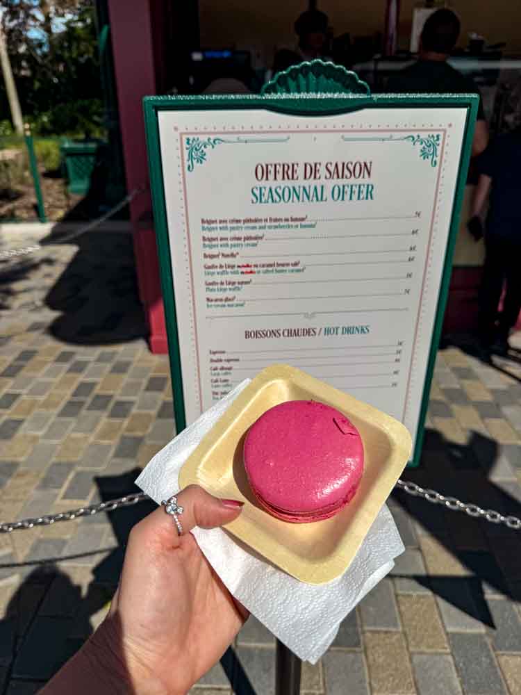 Bright pink raspberry ice cream macaron sandwich held in front of a seasonal snack menu at Disney Adventure World.