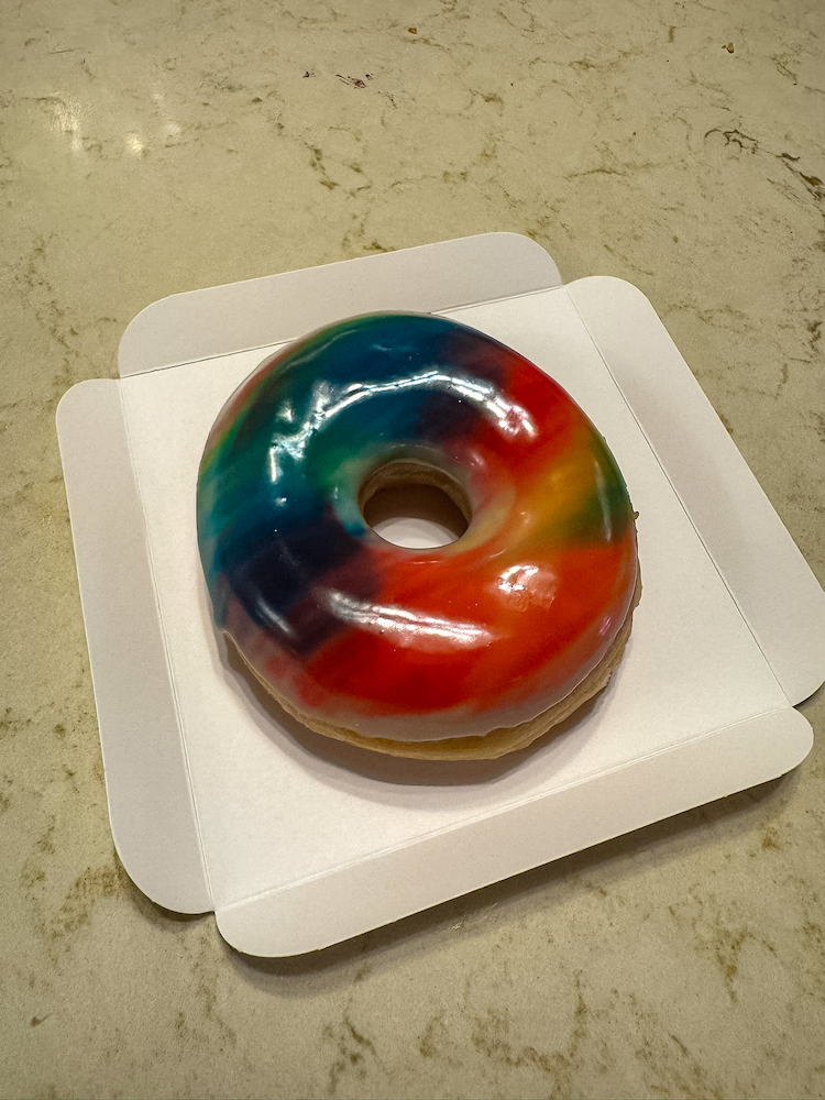 Round Disney donut covered in glossy rainbow-coloured icing, displayed in a white cardboard tray; rates one of the Best Snacks at Disneyland Paris in 2026 for sweet snacks