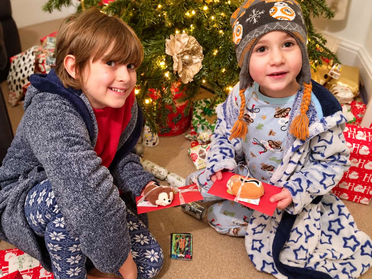 Two young boys in dressing gowns and pyjamas sit by the Christmas tree on Christmas morning, smiling and holding red envelopes topped with little Chip and Dale toys, part of a surprise Disneyland Paris for Christmas reveal.